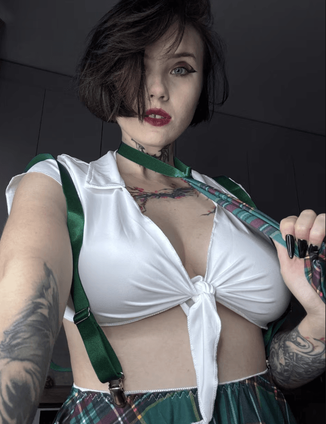 Woman wearing a white tied-front top with green suspenders and a plaid skirt, posing close-up with tattoos visible on shoulders and neck.