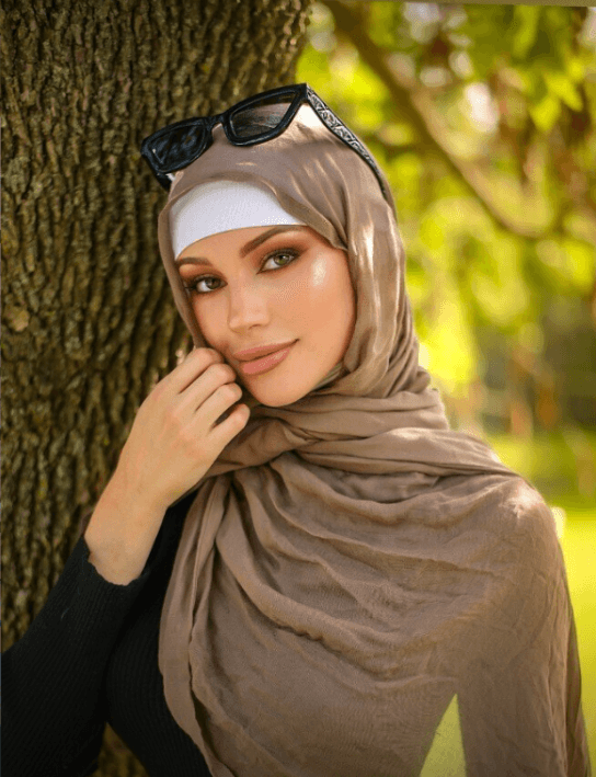 Woman wearing a beige hijab and white headband, leaning against a tree in a sunlit park.