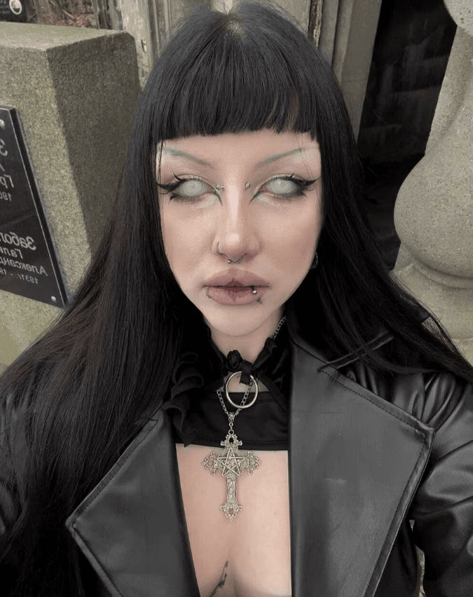 Person with long black hair and blunt bangs, gothic makeup, facial piercings, wearing a black leather jacket and a large ornate cross necklace.