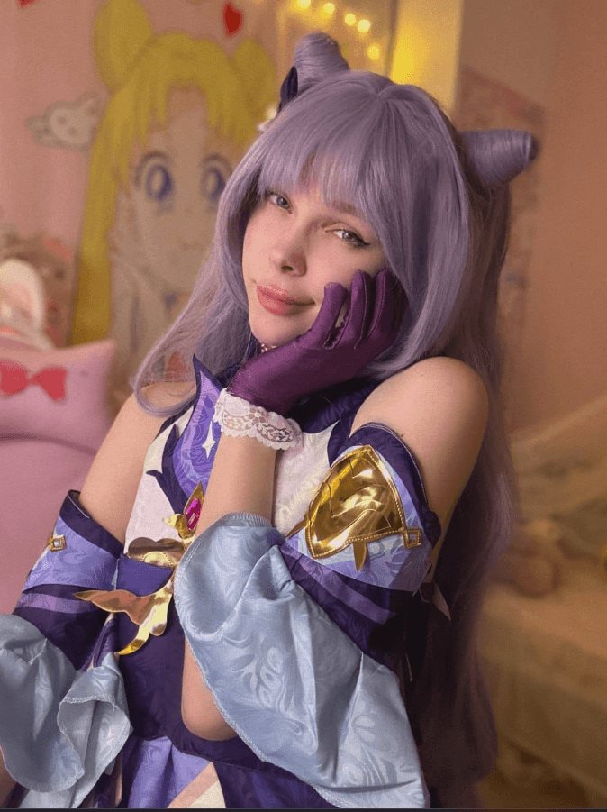 Cosplayer with long lavender wig and cat-ear headband, wearing a purple and blue fantasy costume, resting chin on gloved hand in a pastel room