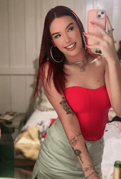 Self-portrait of a woman with red hair taking a mirror selfie, wearing a red strapless top and green skirt, showing tattoos on her arm and hoop earrings, holding a pink phone.
