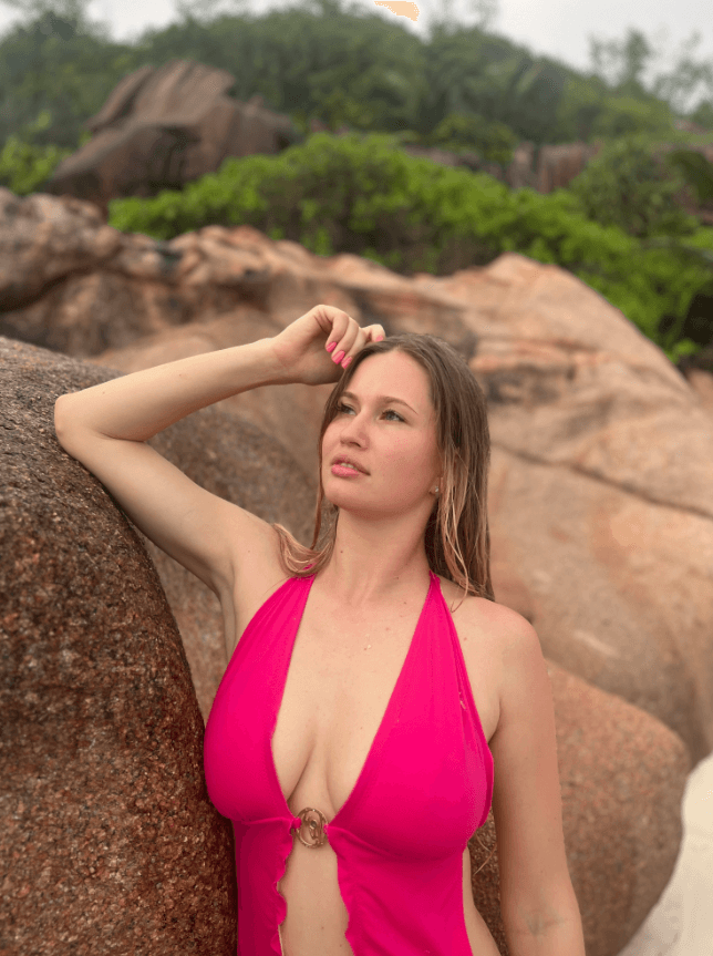 Woman in a bright pink halter swimsuit leaning against a large rock on a rocky beach with greenery in the background, gazing into the distance.