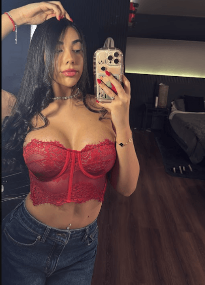 Selfie of a woman in a red lace bustier taking a mirror photo in a bedroom, holding a gold phone with painted nails.