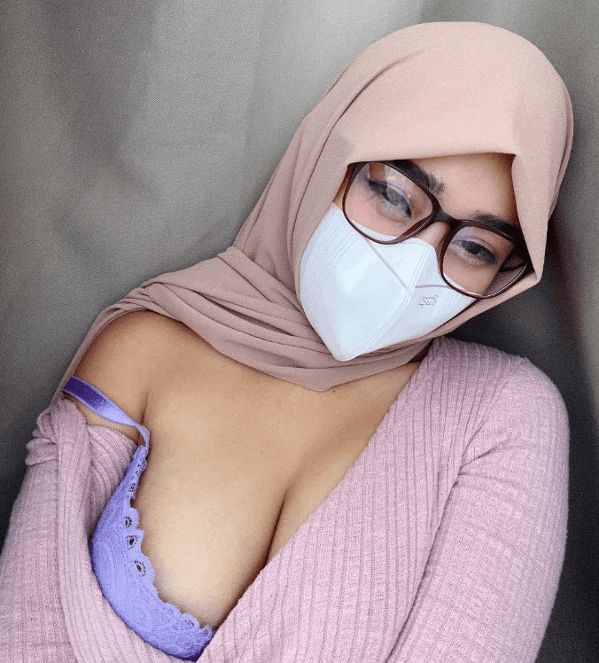 Woman wearing a light pink hijab, glasses, and a white mask, with a pink knit top and lavender lace bra showing at the neckline.