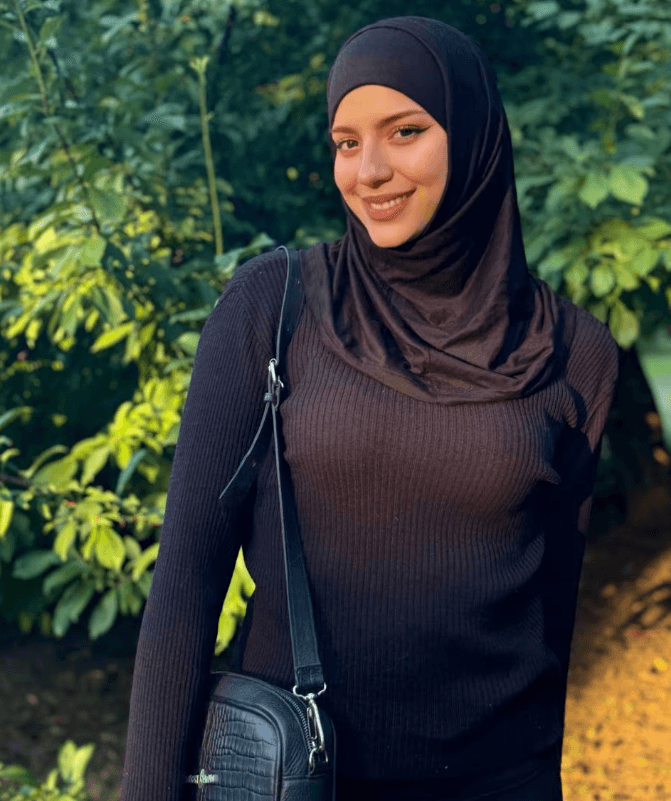 Woman wearing a black hijab and brown knit top, outdoors with green foliage, carrying a black handbag over the shoulder.