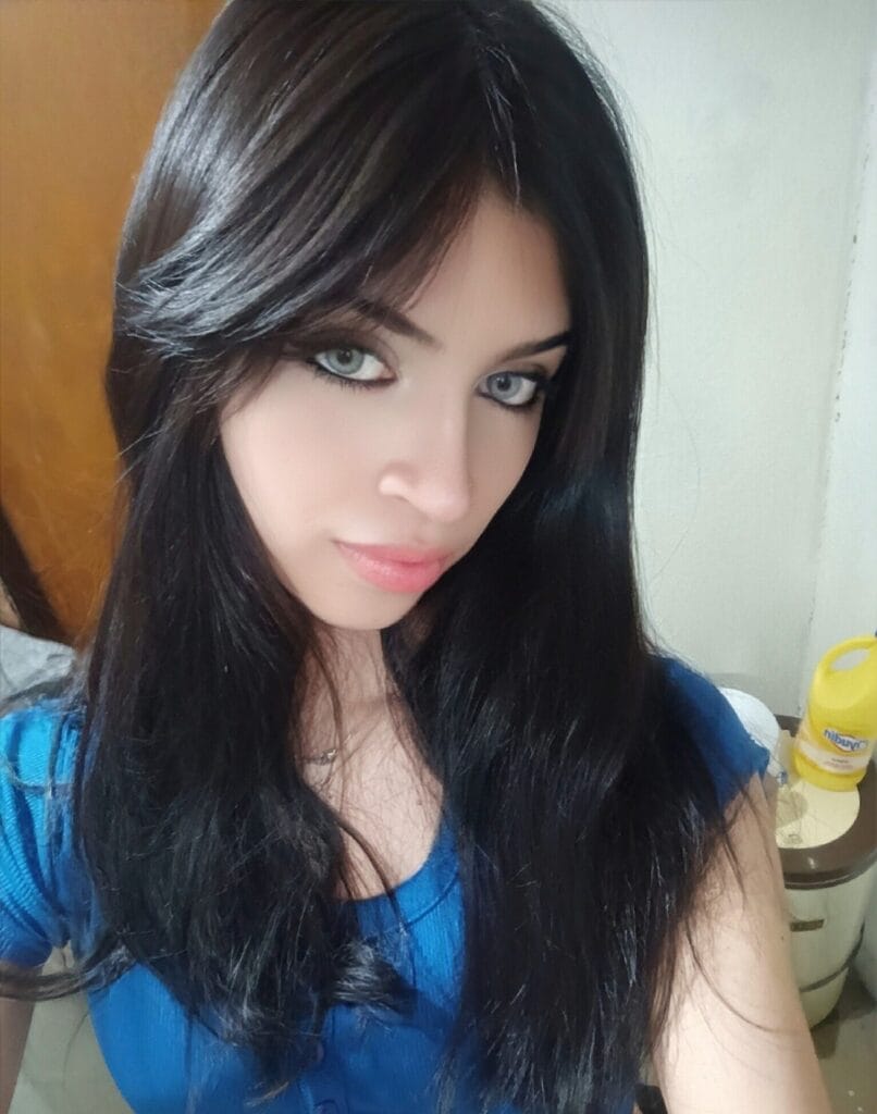 Close-up of a woman with long dark hair and blue eyes, wearing a blue top, looking at the camera indoors with a yellow bottle in the background.