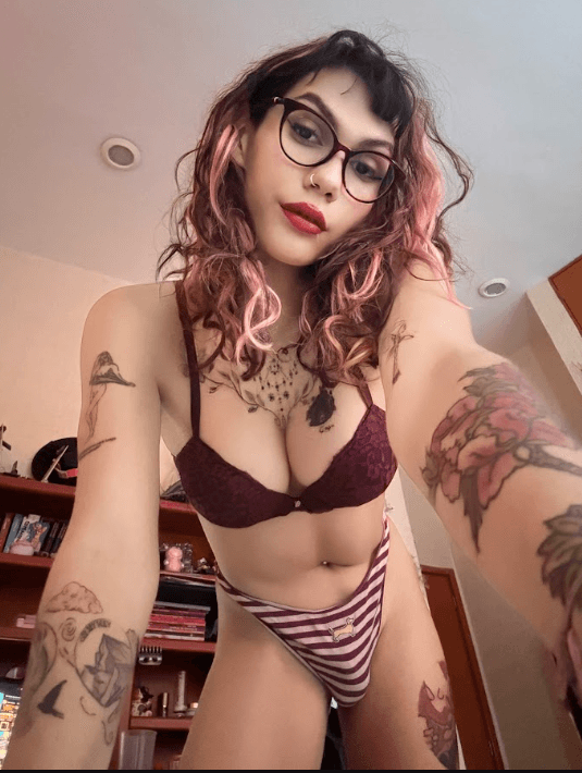 Selfie of a tattooed person with pink-dyed hair and glasses wearing burgundy lace bra and striped panties, in a room with shelves in the background.