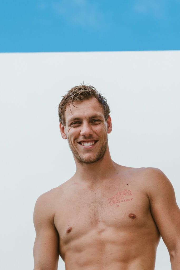 Smiling shirtless man with short hair against a white wall and blue sky; tattoo on his left chest reads 'survivor'