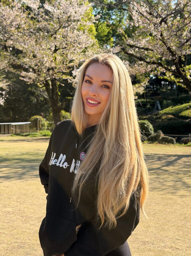 Smiling woman with long blonde hair standing in a sunny park with blooming cherry blossoms behind her.