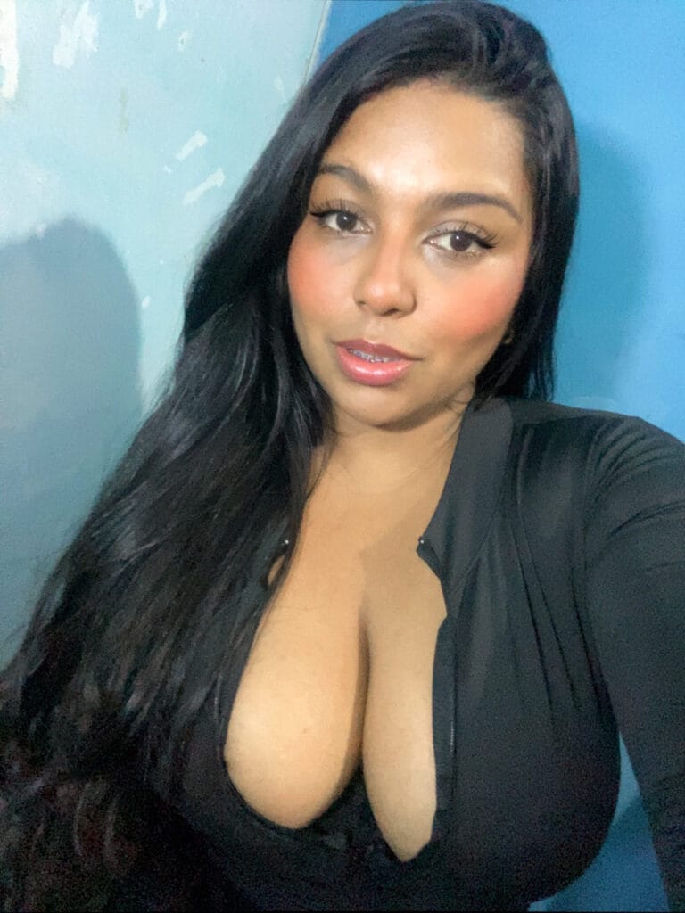 Close-up portrait of a woman with long black hair, wearing a black top, smiling softly at the camera against a blue wall background.