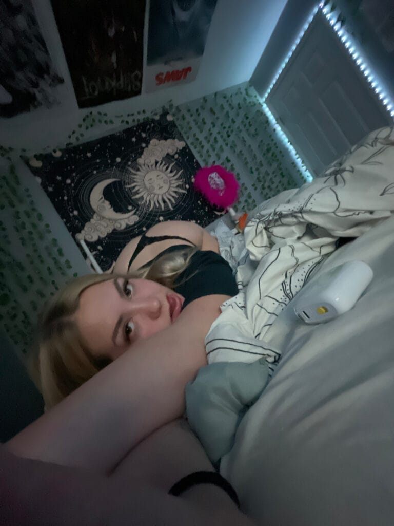 Person lying on a bed in a dimly lit room, facing the camera with one arm overhead, wearing a black top and surrounded by patterned bedding and wall decor including posters and string lights.