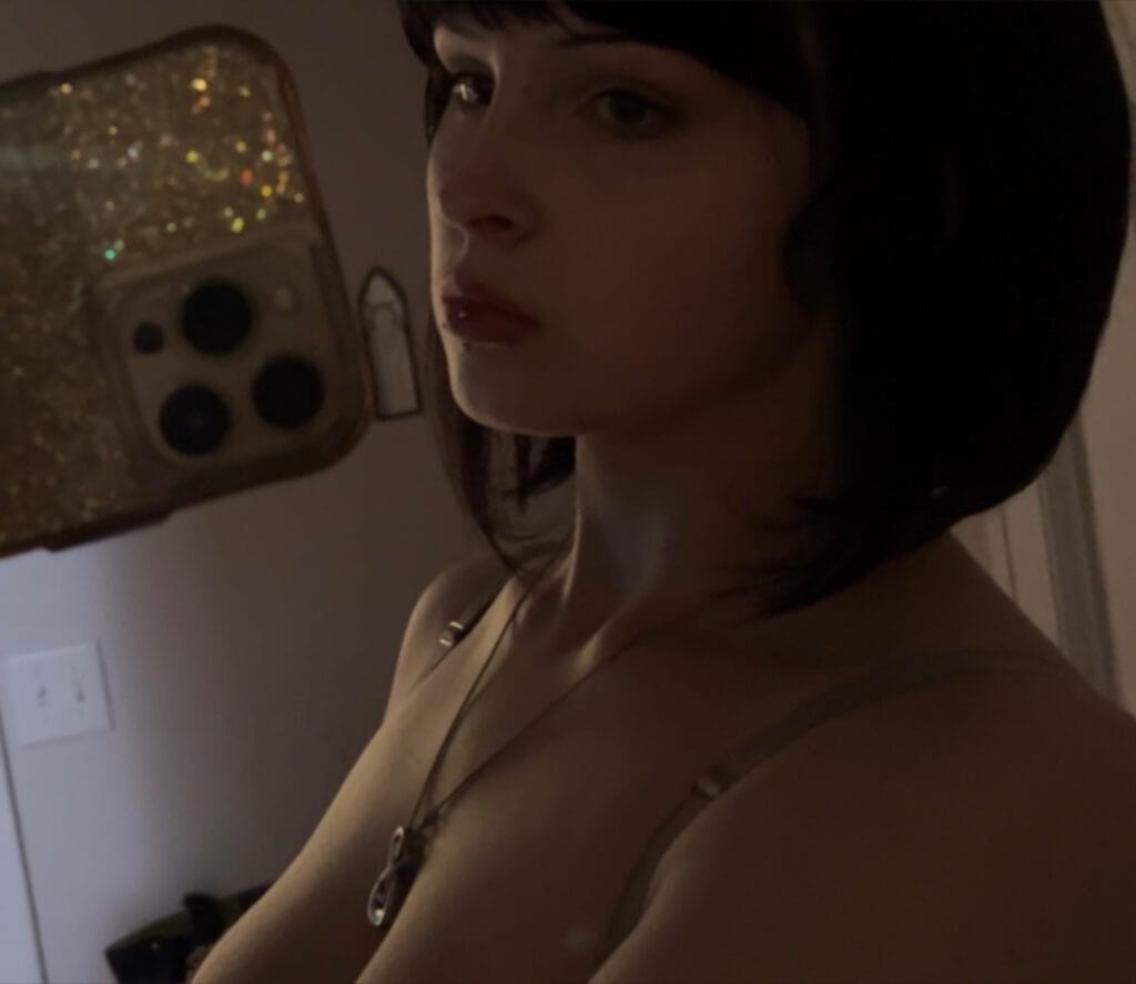Young person in a dim room wears a hoodie and necklace, gazing to the side while holding a glittery smartphone with a patterned case (mirror selfie).