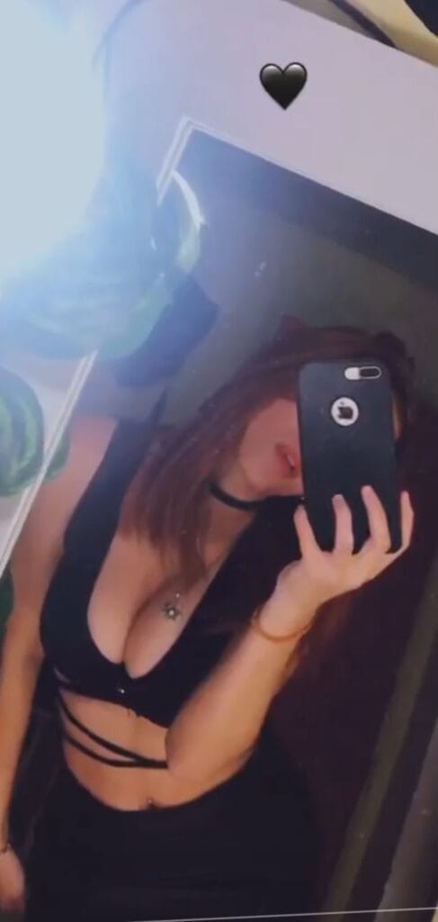 Mirror selfie of a person wearing a black strappy bralette and choker, holding a phone to take the photo; a black heart sticker is above.