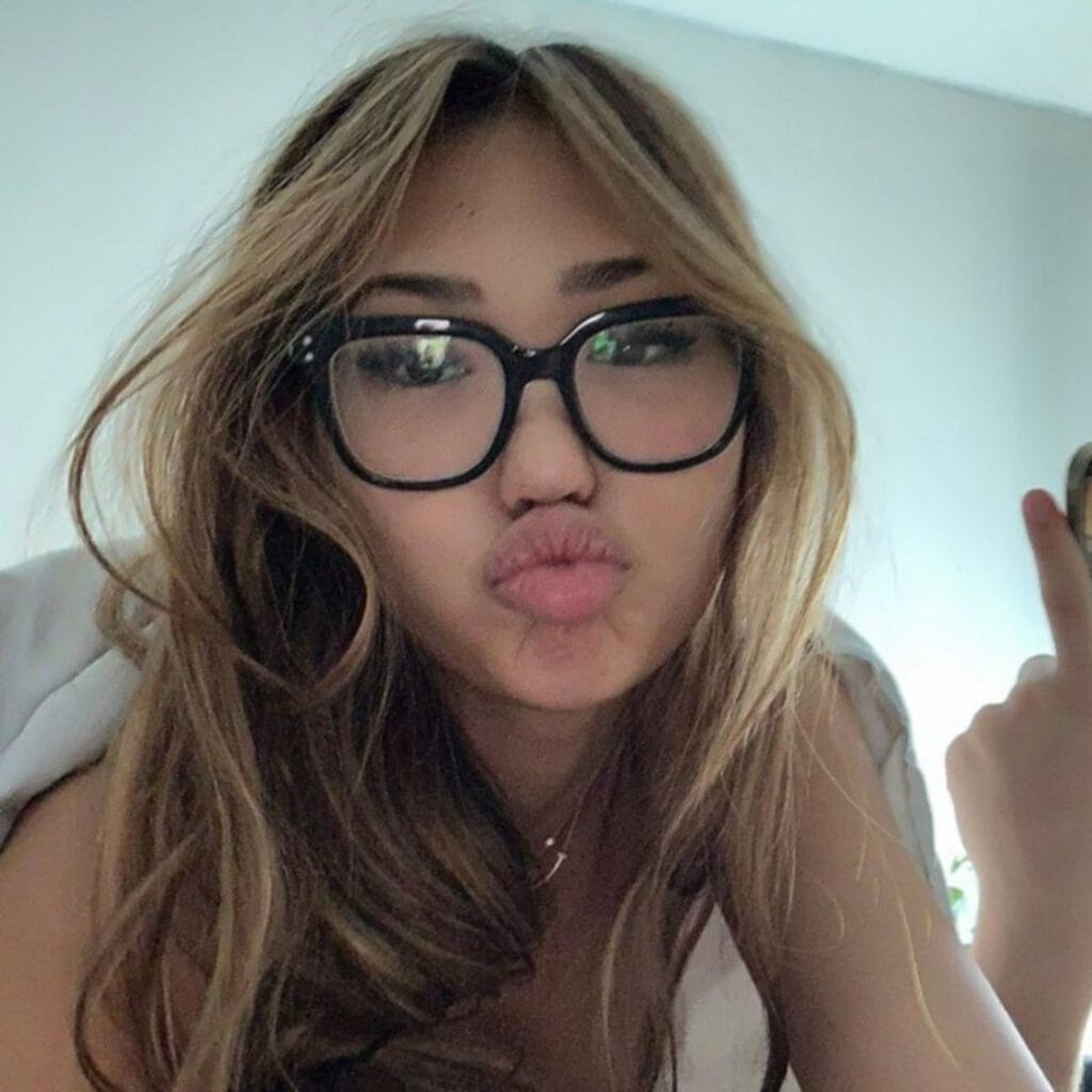 Close-up of a person with long light-brown hair and black glasses, lips puckered as if blowing a kiss.