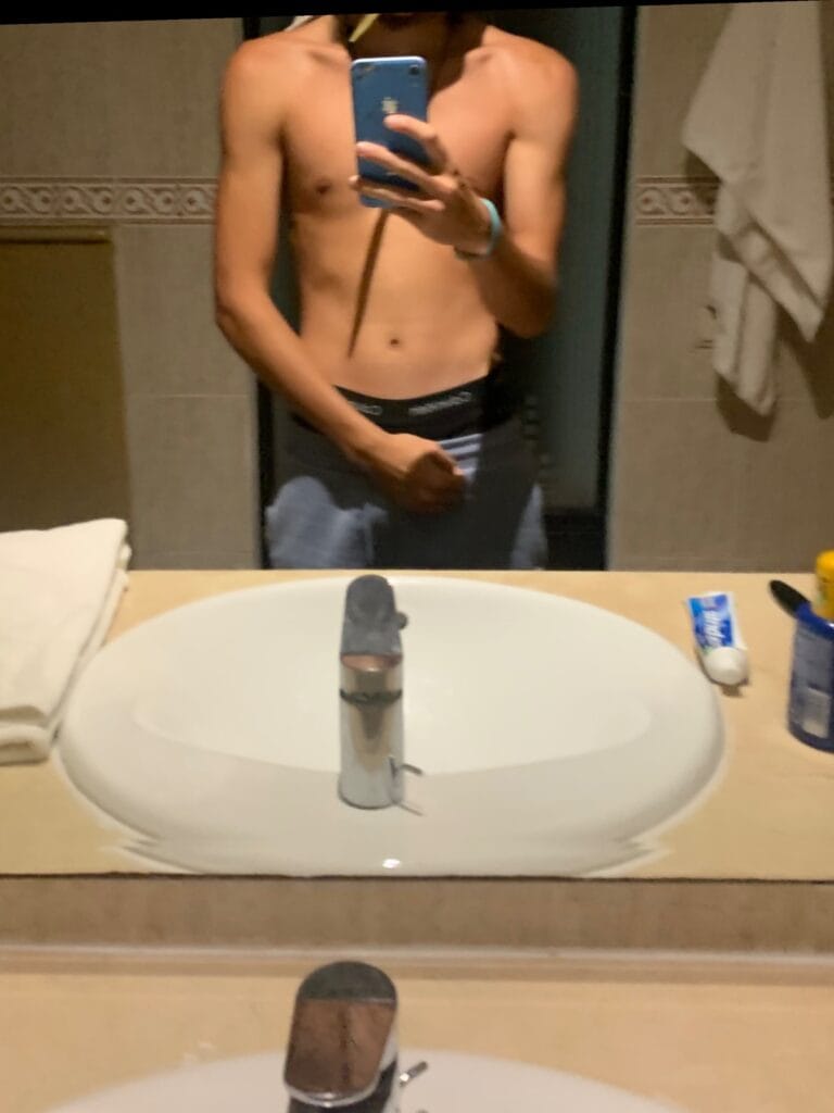 Shirtless person taking a mirror selfie in a bathroom, holding a blue smartphone, wearing dark shorts with underwear waistband visible at the top, sink and towels in the background