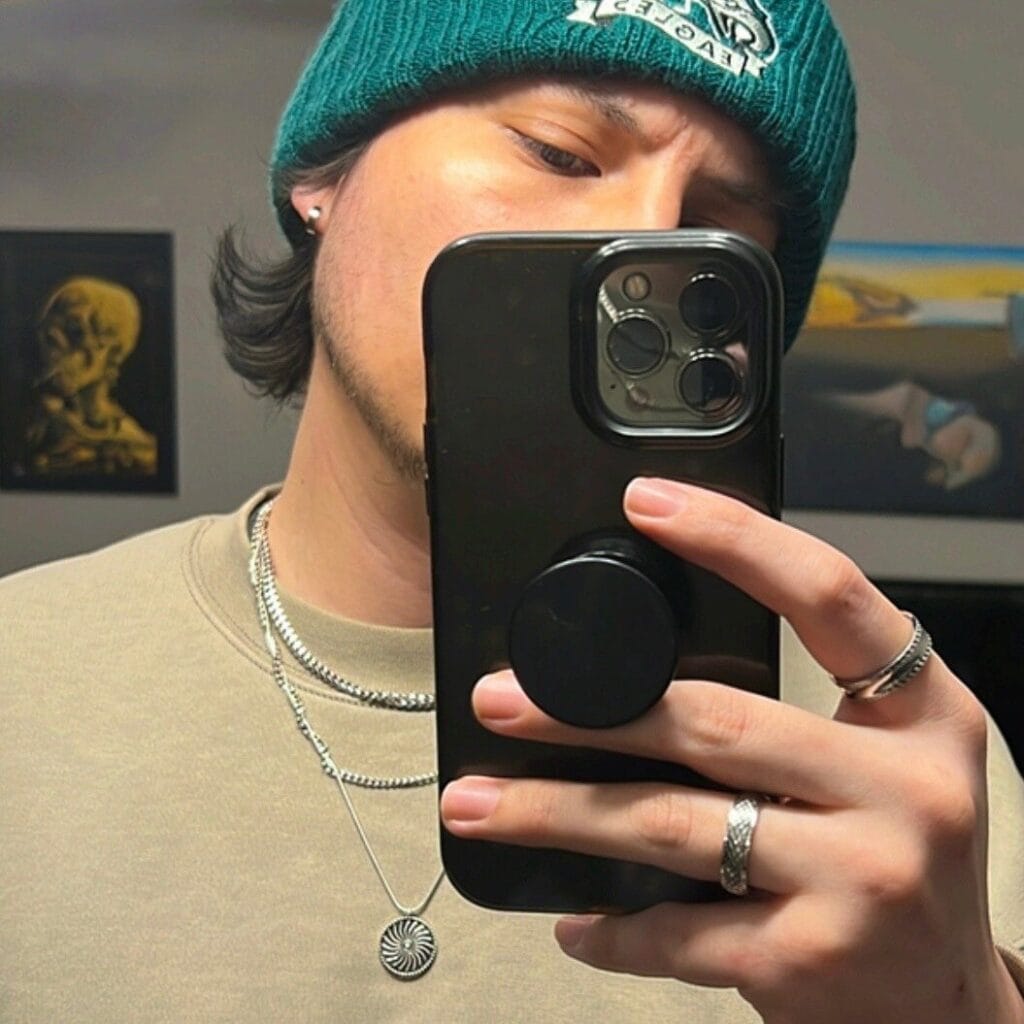 Person takes a mirror selfie holding a black phone; teal beanie, beige shirt, and layered jewelry visible.