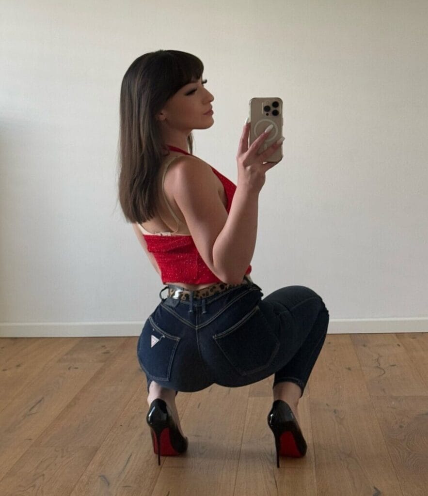 Side-profile selfie of a woman in a red crop top and dark jeans, squatting and holding a phone in a mirror.