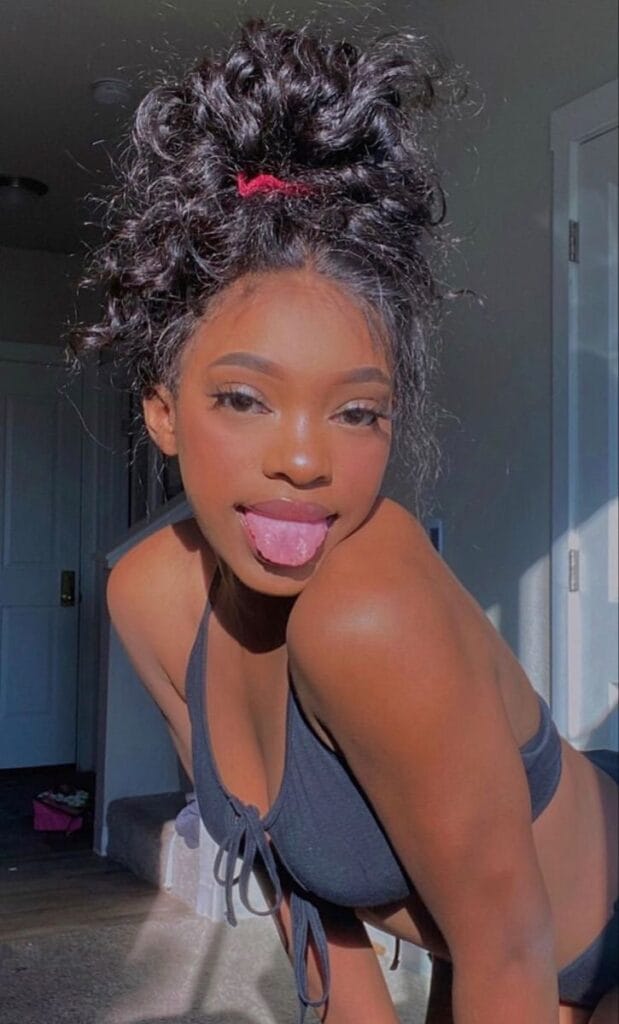 Young woman wearing a dark bikini top leans forward in sunlight, sticking out her tongue playfully with a messy bun hairstyle.