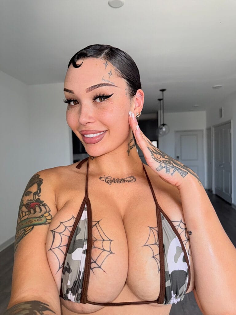 Smiling woman with slicked-back hair and multiple tattoos posing in a camouflage bikini top indoors.