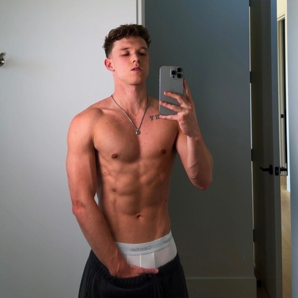 Young man shirtless in a mirror selfie, wearing white Calvin Klein waistband and a necklace, flexing his upper body in a modern room.