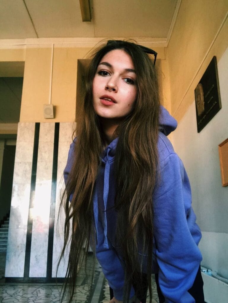 Young woman with long brown hair in a blue hoodie leaning toward the camera in an indoor hallway.