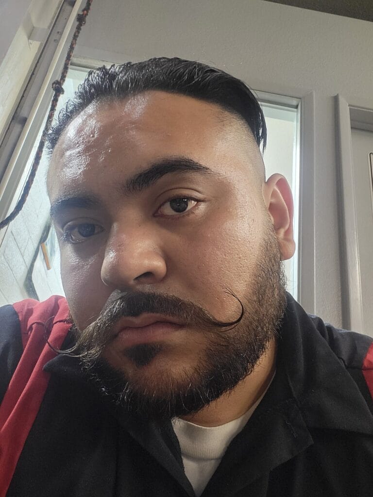 Close-up of a man's face with a slicked-back hairstyle, beard, and mustache, indoors near a window.