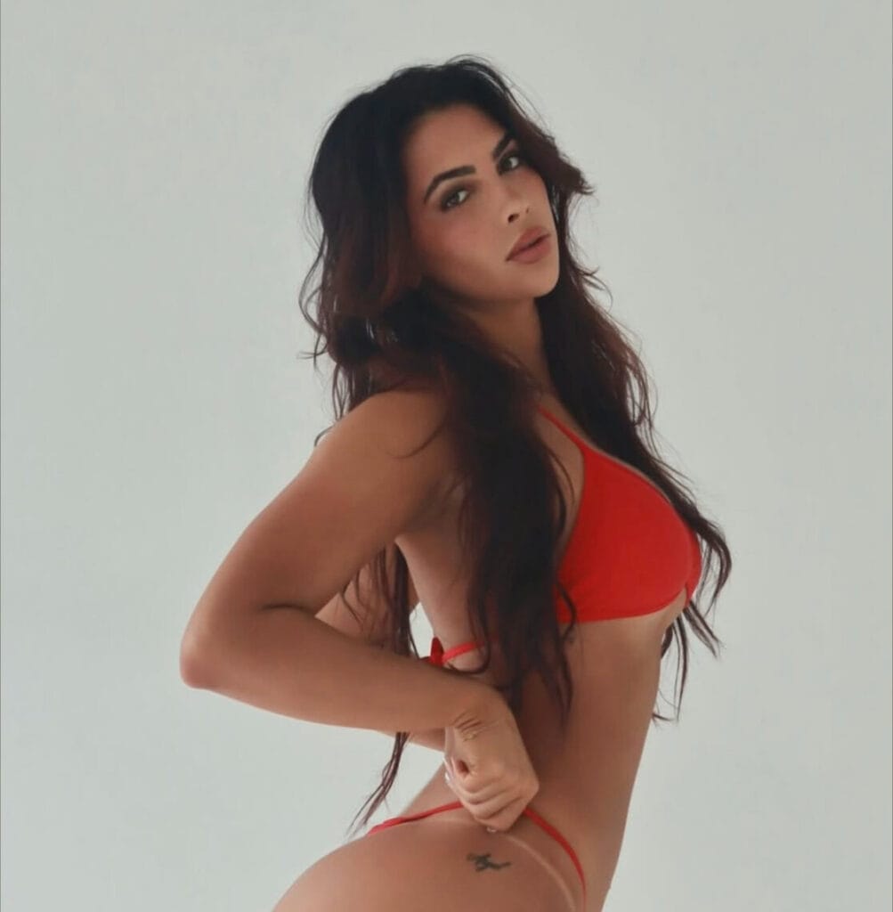 Woman with long dark hair wearing a red bikini, posed in profile against a light background, looking at the camera.