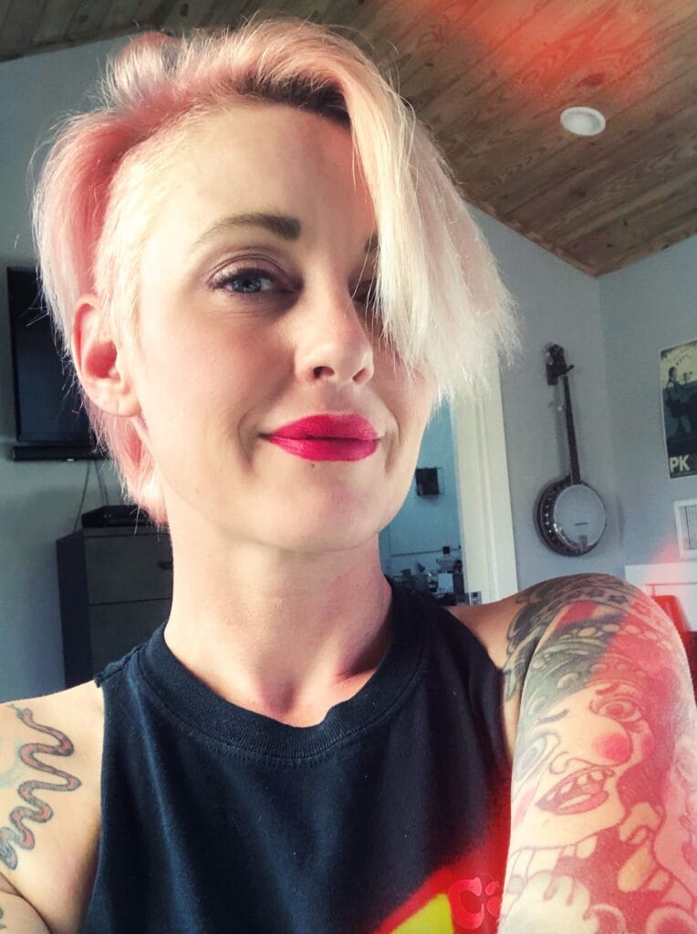 Self-portrait of a person with short pink-blonde hair, pink lipstick, and visible tattoos on arms in a room with wood ceiling and light.