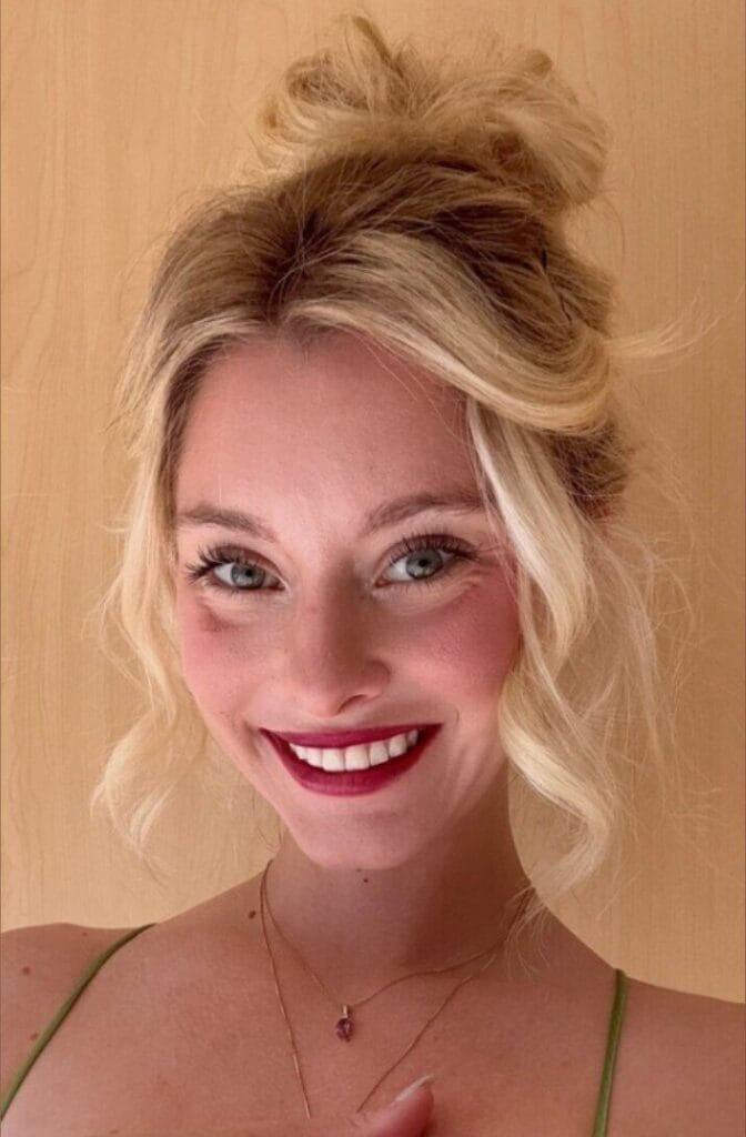 Close-up of a smiling young woman with blonde hair in a messy bun, wearing red lipstick and a delicate necklace against a light wooden background.