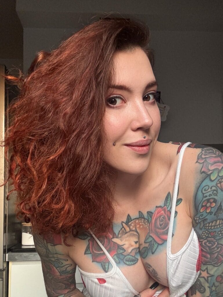 Close-up portrait of a woman with long, curly red hair and colorful floral tattoos on her arm and chest, wearing a white striped top.