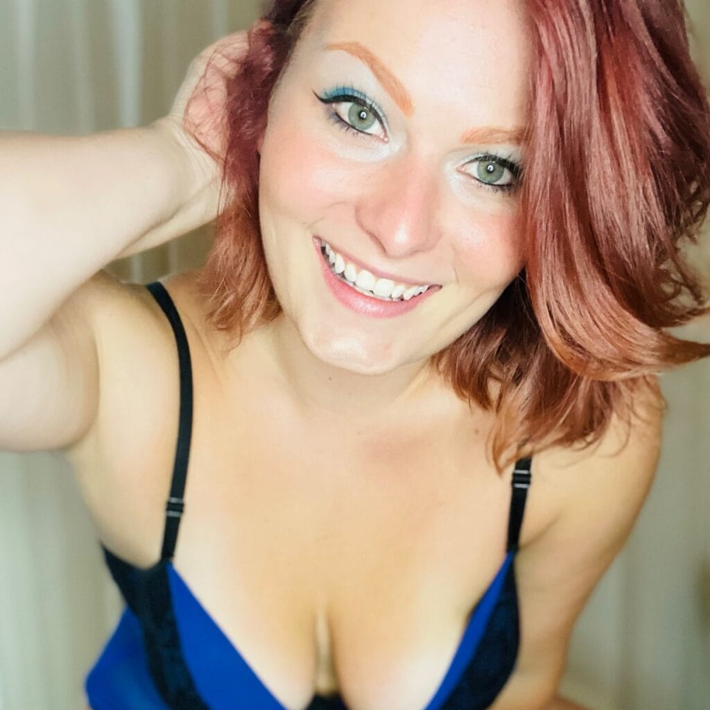 Close-up selfie of a smiling woman with red hair, green eyes, wearing a blue top with black straps.