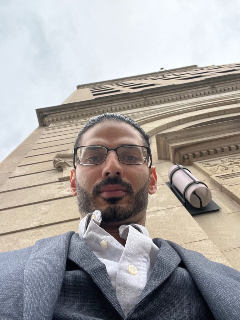 Self-portrait of a man with glasses and a gray suit, taken from a low angle against a tall stone building with decorative molding in the background