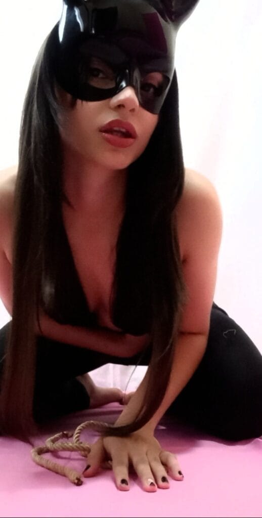 Woman wearing a black cat mask, kneeling on a pink surface with long dark hair and a rope nearby.