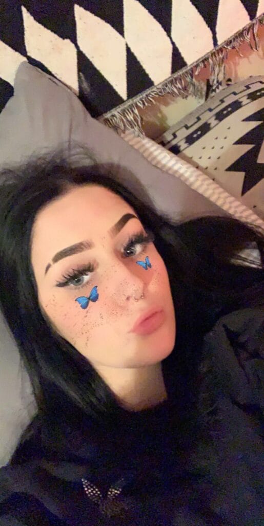 Person lying on a patterned blanket, dark hair, bold brows, freckles, and blue butterfly filter on cheeks.