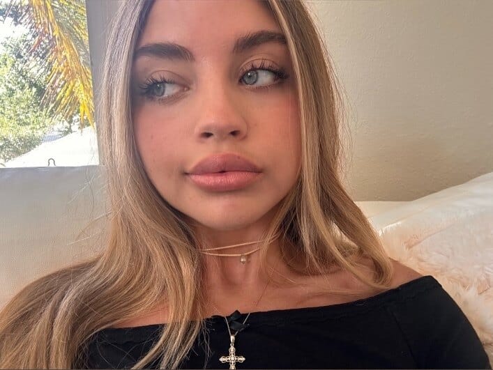 Close-up portrait of a young woman with blonde hair and blue eyes, looking to the left, wearing a black off-the-shoulder top and a cross necklace.