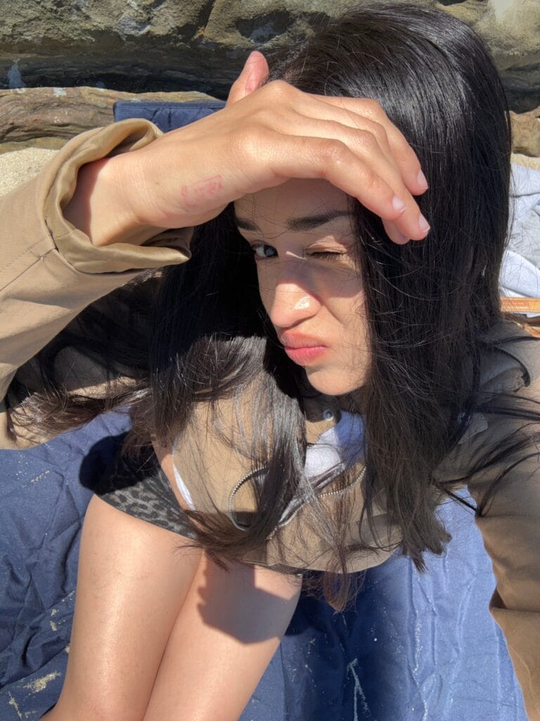 Person lying on rocky ground shading eyes with forearm, sunlit scene, wearing beige jacket and dark hair