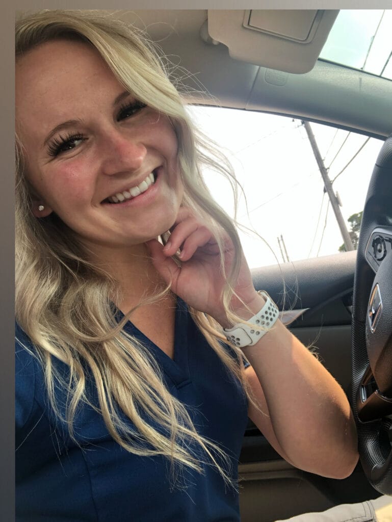 Smiling blonde woman in a navy top sits in a car, hand near her chin, wearing a white perforated smartwatch.