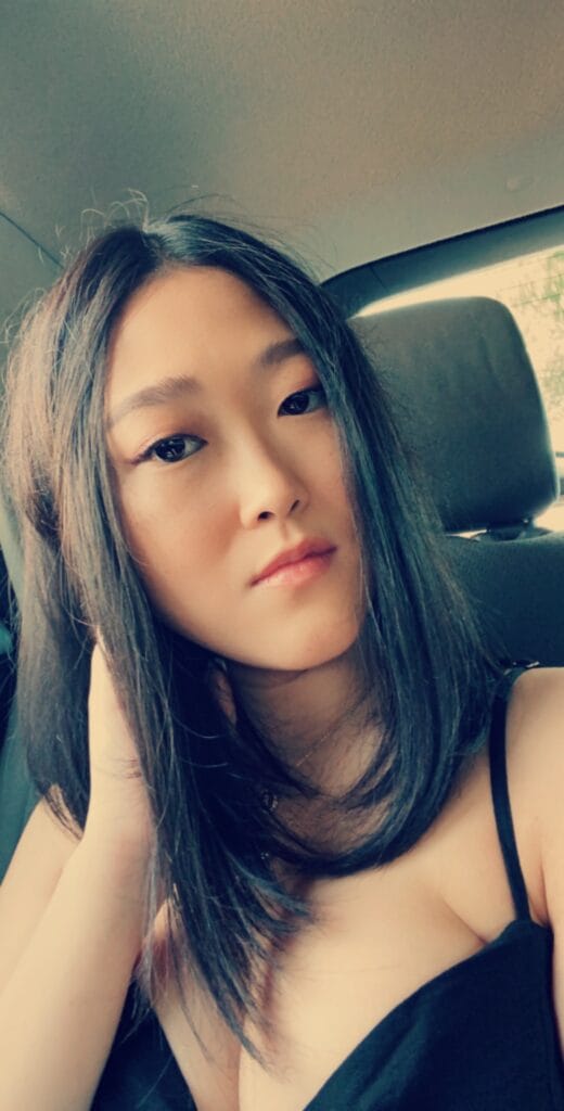 Portrait of a young woman with shoulder-length dark hair, looking at the camera while seated in a car, wearing a black top.