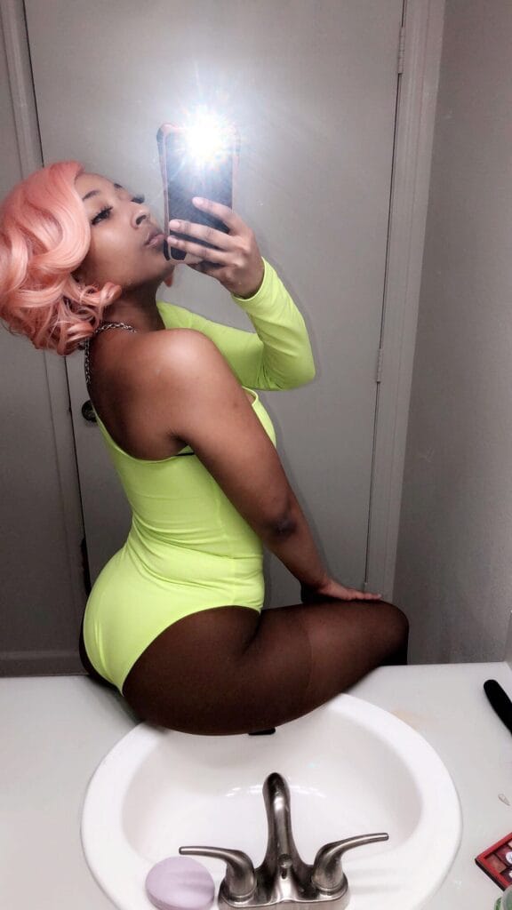 Person with pink hair in a neon lime swimsuit taking a mirror selfie in a bathroom with the phone flash visible in the reflection.