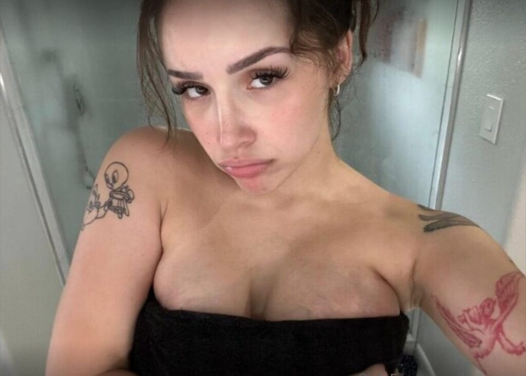 Close-up portrait of a topless woman wearing a black strapless top, with tattoos on her left arm, looking at the camera.