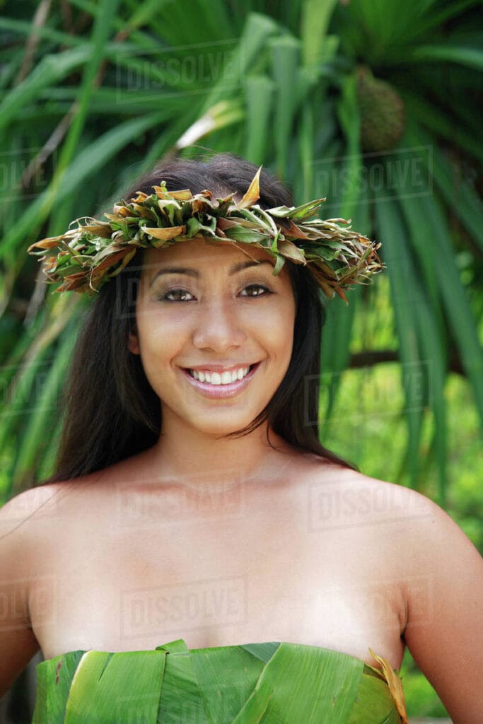 Smiling woman wearing a leafy crown outdoors, with tropical greenery in the background.