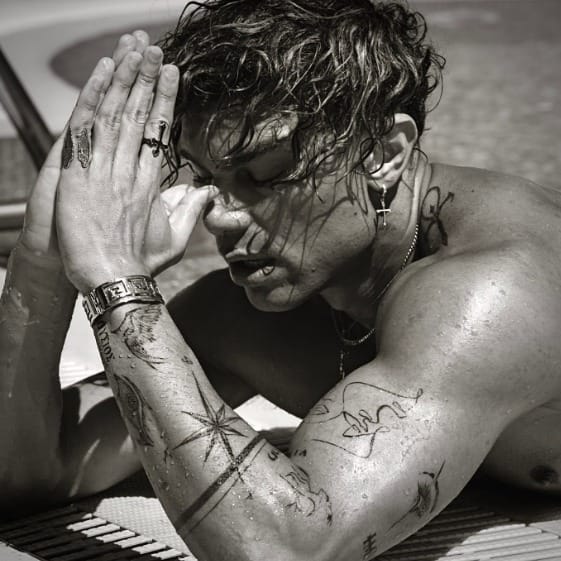Black-and-white photo of a shirtless man with multiple tattoos, hands pressed together near his face in a prayer-like pose.
