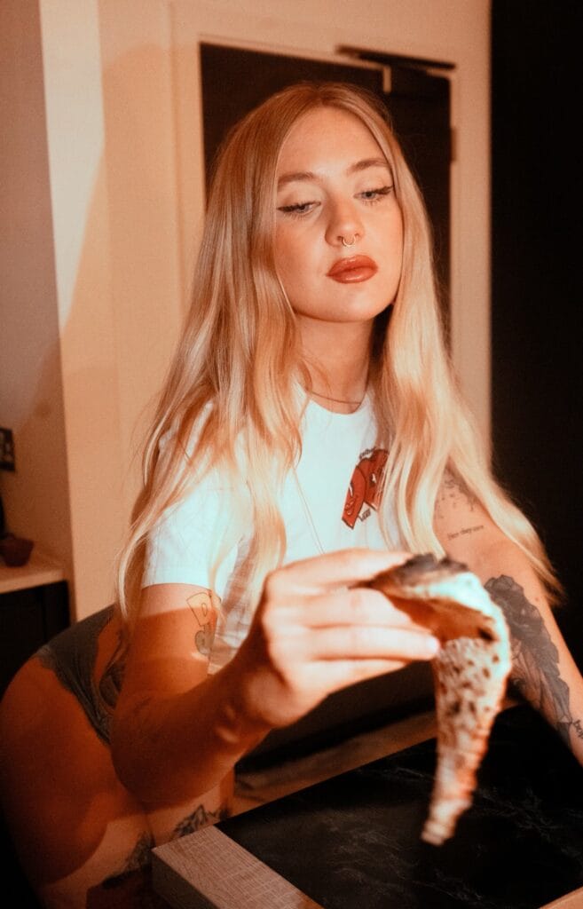 Young woman with long blonde hair and a septum piercing holds and licks a cone with blue ice cream indoors, tattoos visible on her arm.