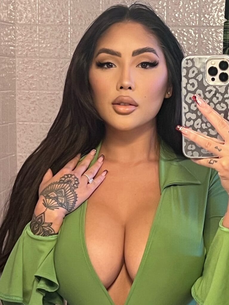Self-portrait of a woman with long dark hair wearing a low-cut green top, showing a bold makeup look and a floral tattoo on her left forearm while taking a mirror selfie with a patterned phone case.