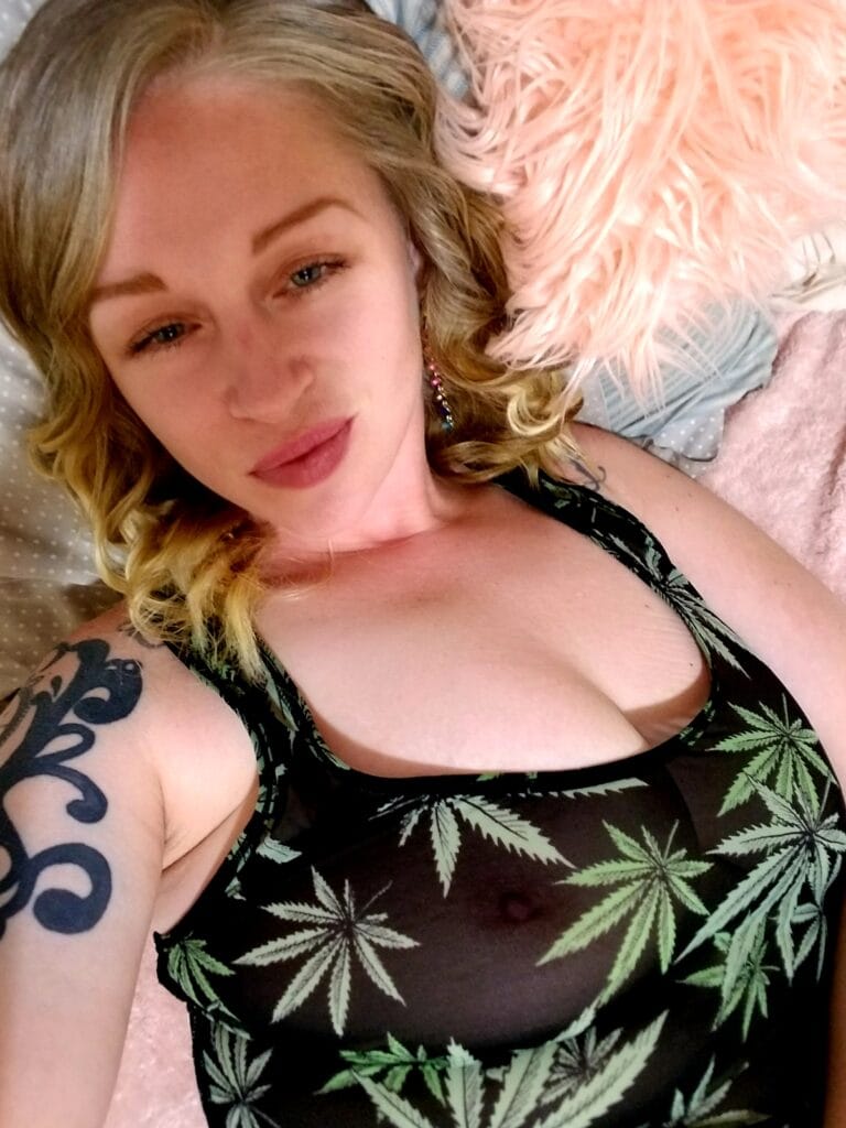 Woman lying down, resting on a pink fuzzy blanket, wearing a black swimsuit top with green marijuana leaf print and visible arm tattoo on the left.