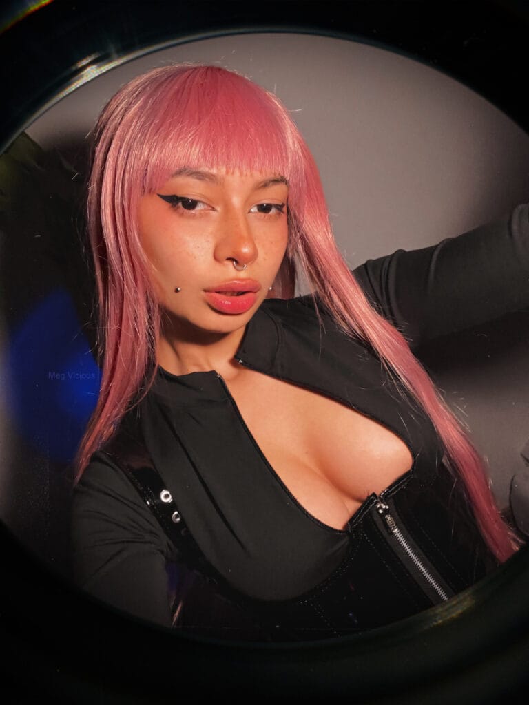 Close-up of a person with pink hair, bold winged eyeliner, septum piercing, and a black zip-up jacket.