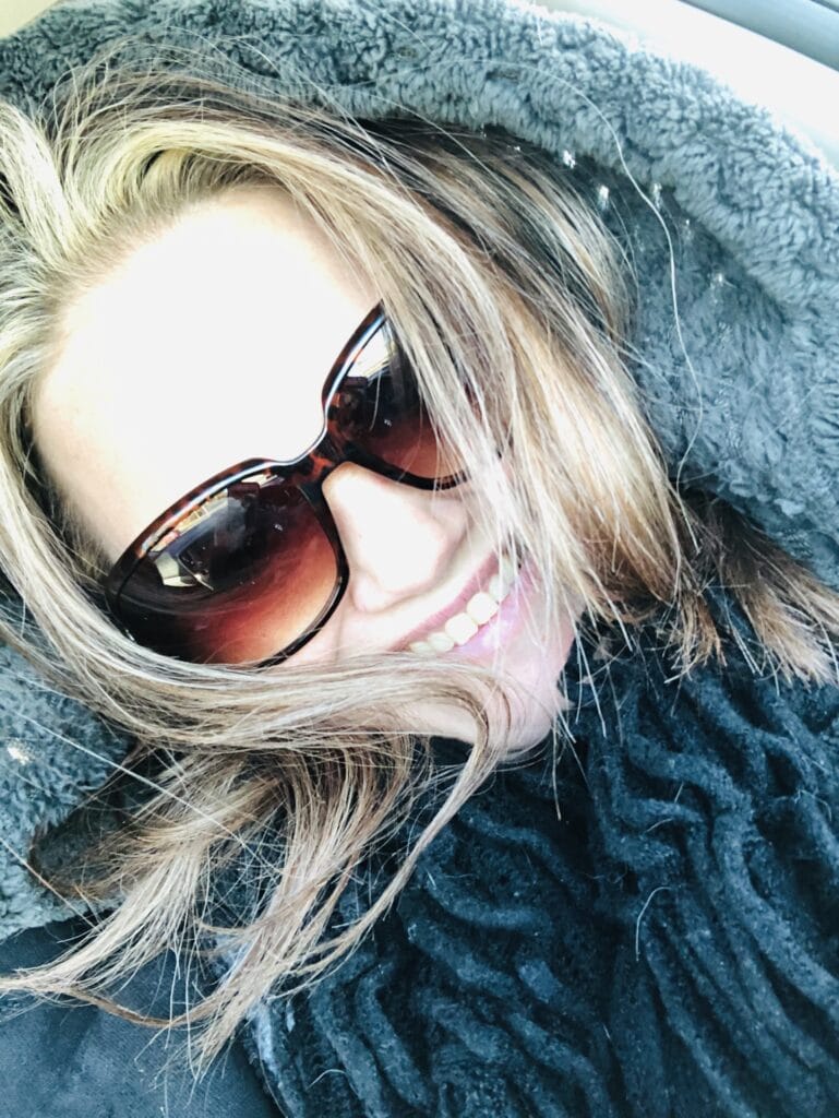 Smiling person wearing oversized brown sunglasses with blond hair framing the face, resting on a plush gray surface.