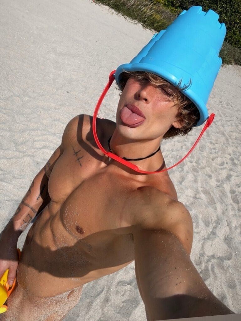 Young man at the beach wearing a blue bucket hat, sticking out his tongue, holding a yellow object in the sand-handed selfie pose.
