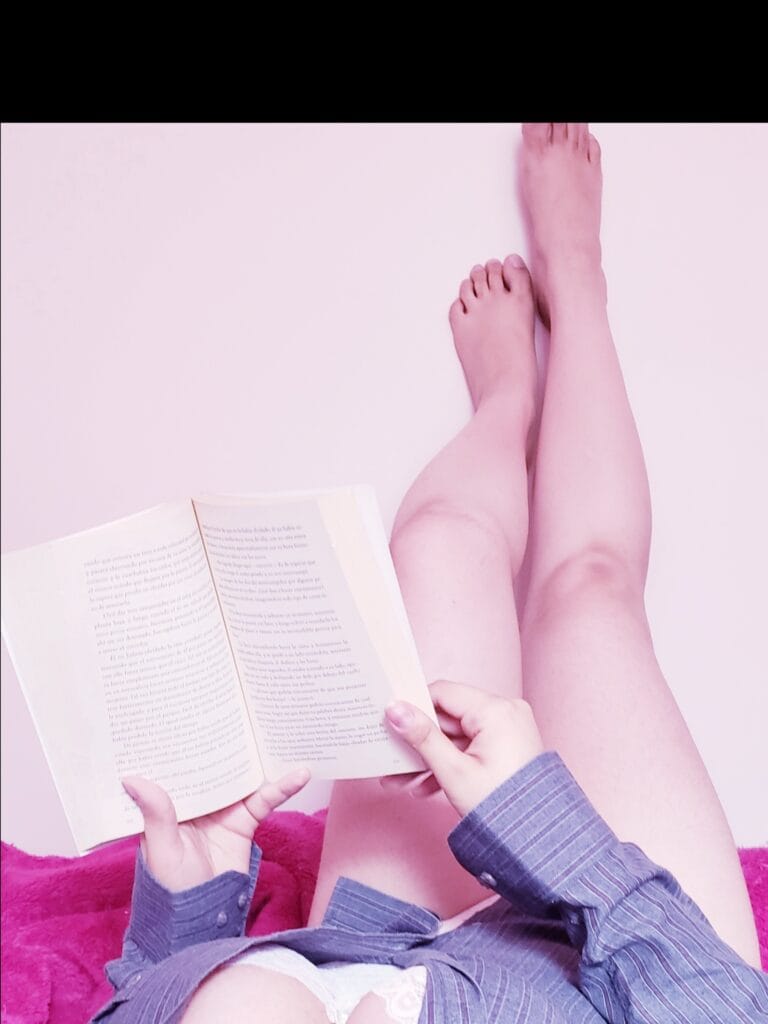 Open book resting on a pink surface with a child’s legs nearby and an adult’s hands turning the pages on the left side.