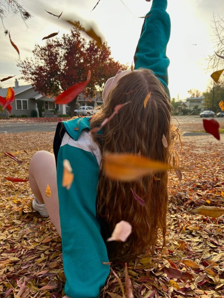 Back view of a person with long brown hair in a teal jacket sitting among a carpet of fallen autumn leaves in a residential street, with one arm raised as leaves swirl around.